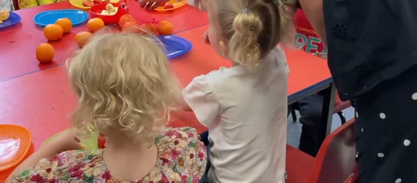 Two young children at snack time at Little Darlings, sitting at a table with fruit and plates
